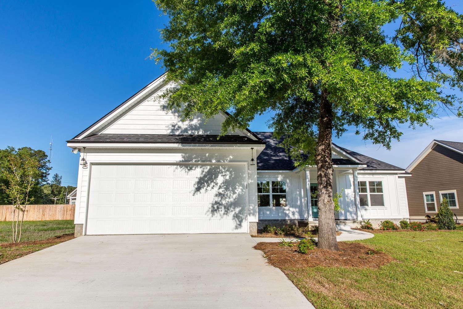 Suburban house with white garage, large tree, and driveway under a clear blue sky