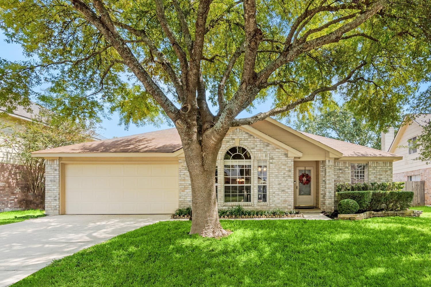 Suburban single-story house with a large front tree, green lawn, and two-car garage