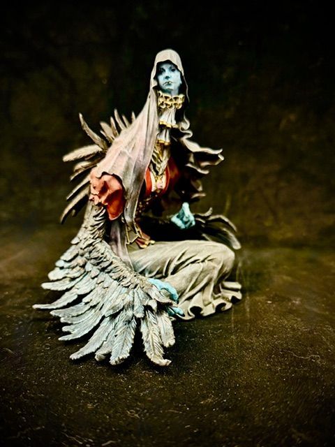 Statue of a seated figure with blue skin and feathered wings, wearing a veil, red and white garments.