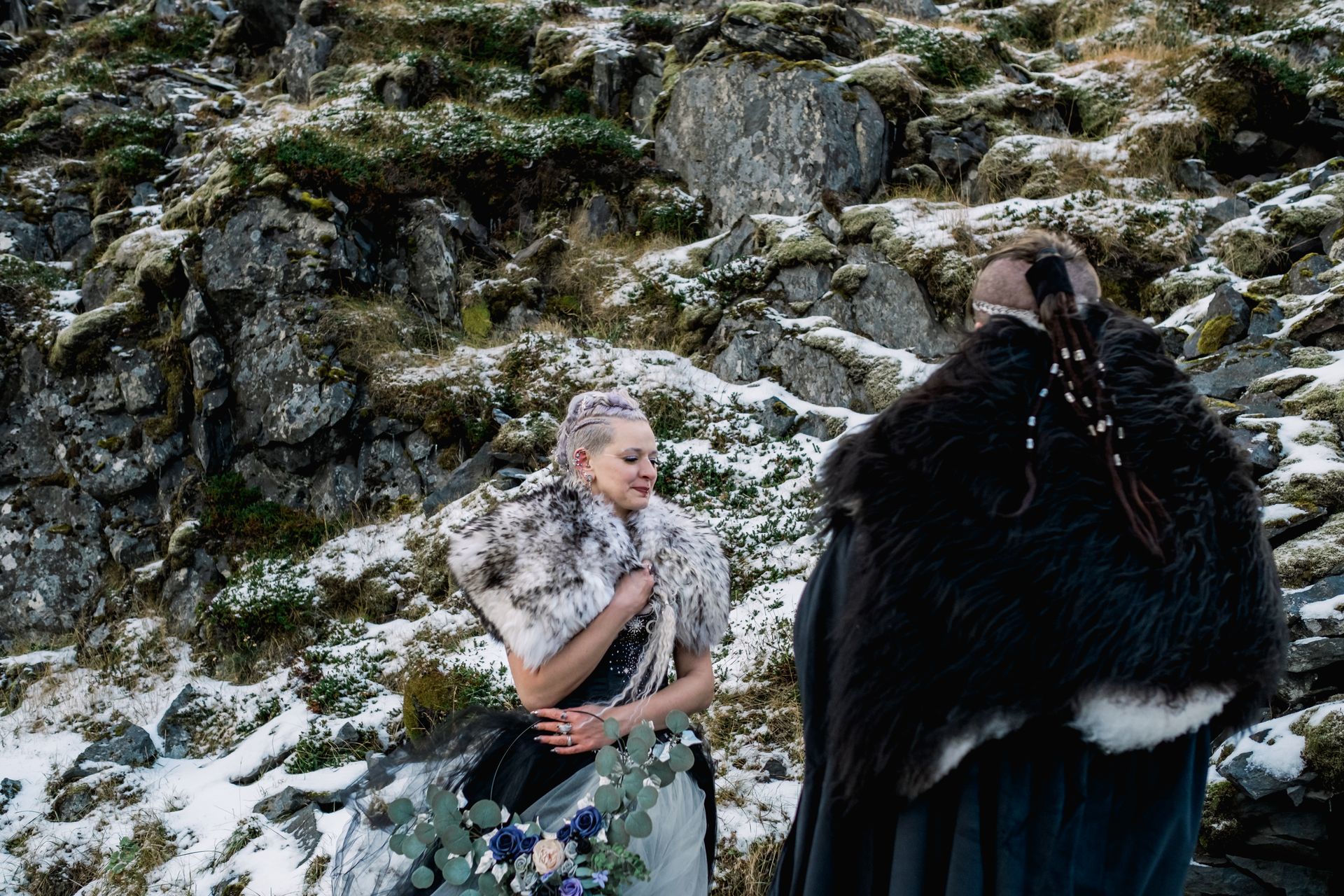 A man in a fur coat is standing next to a woman in a wedding dress in the snow.