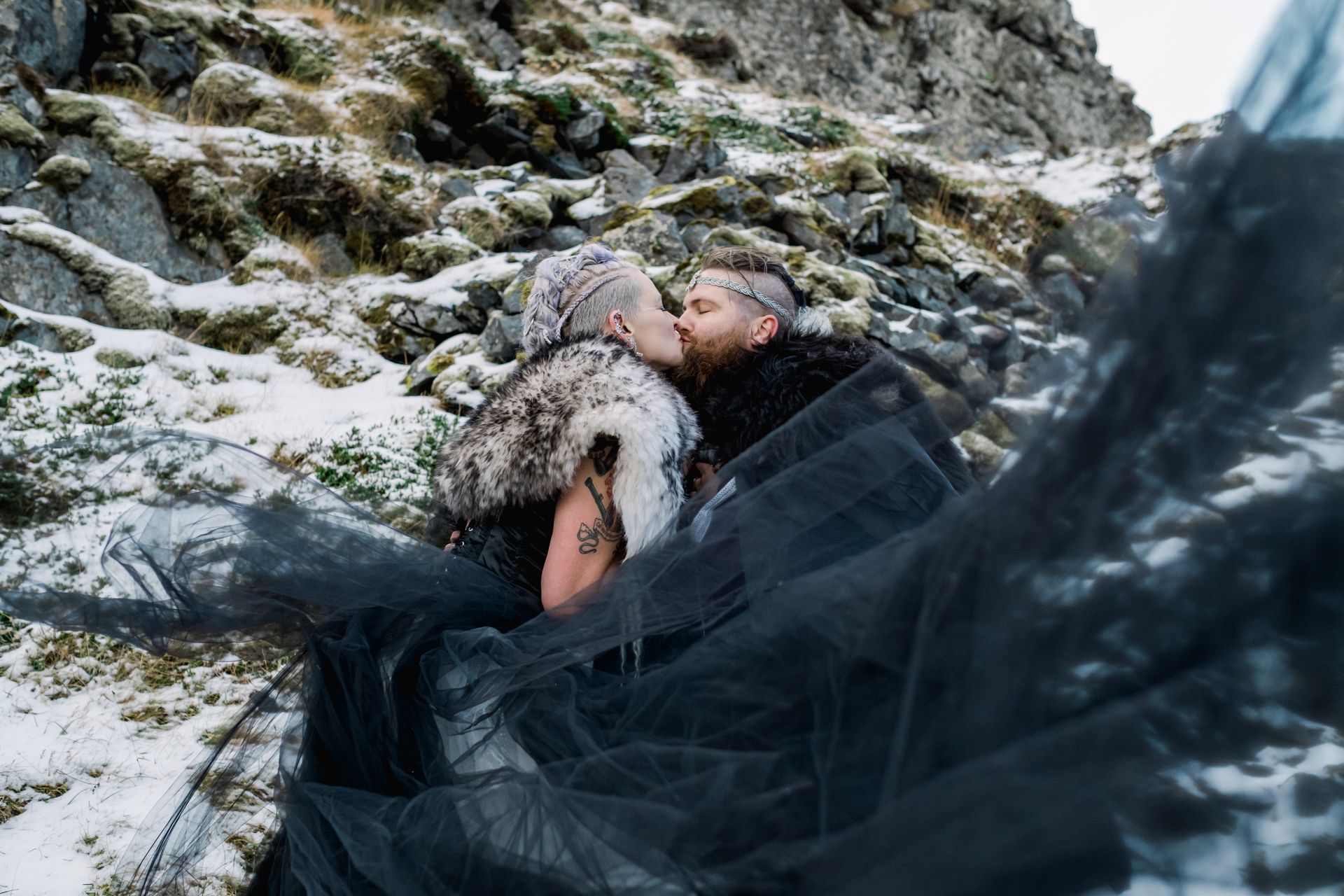A man and a woman are kissing in the snow on top of a mountain.
