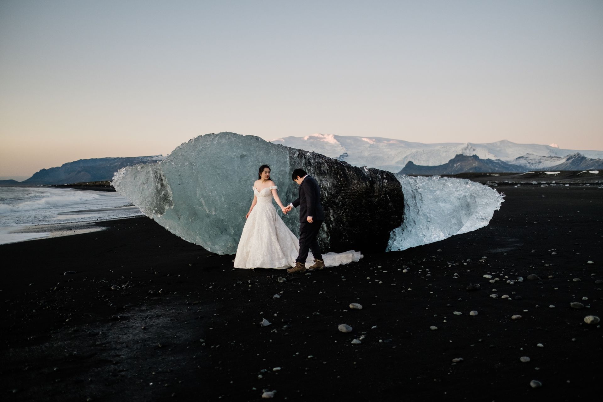 A bride and groom are standing next to a large piece of ice on a beach.