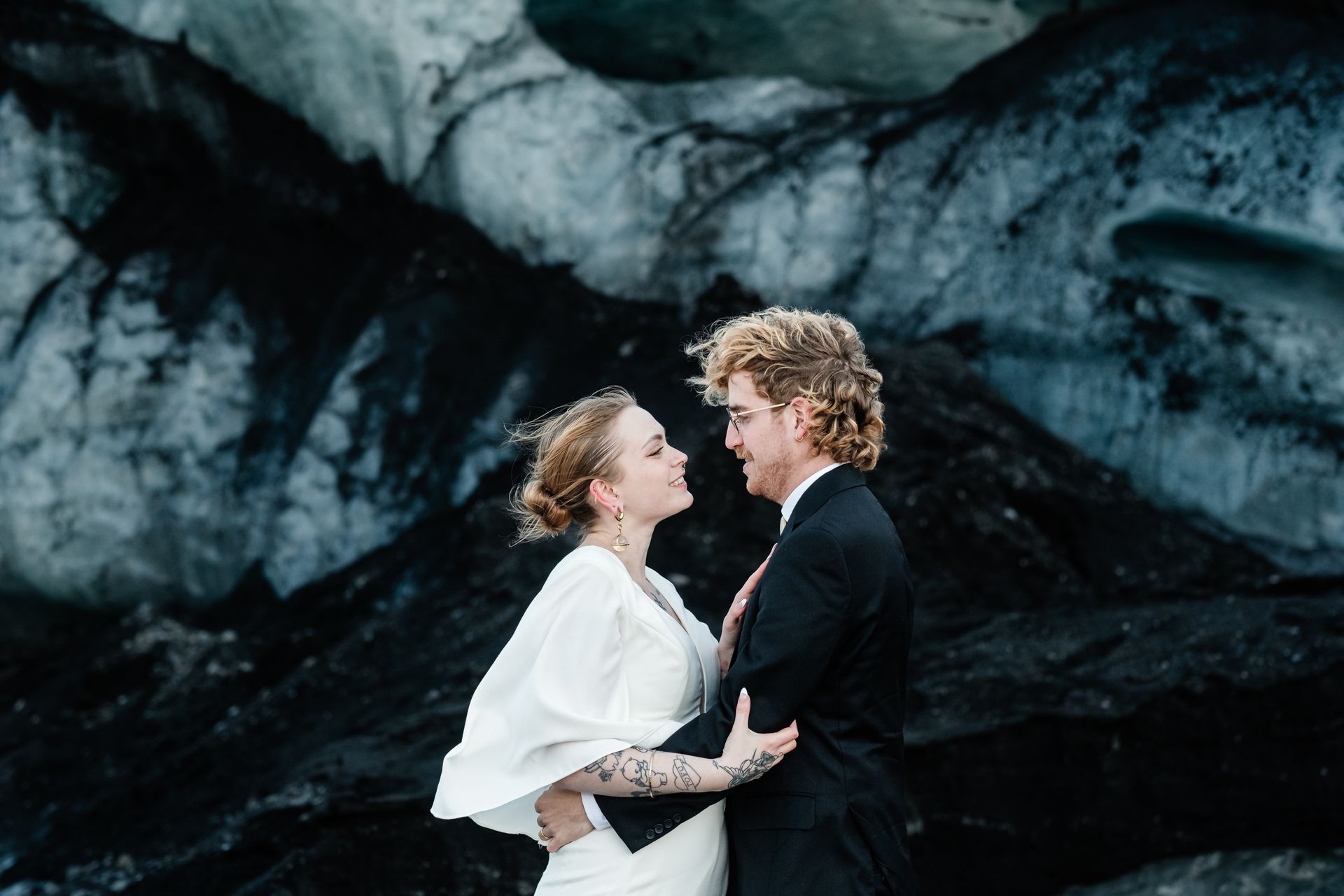 A couple takes stunning elopement photos in Iceland, posing by an ice cave in windy conditions.