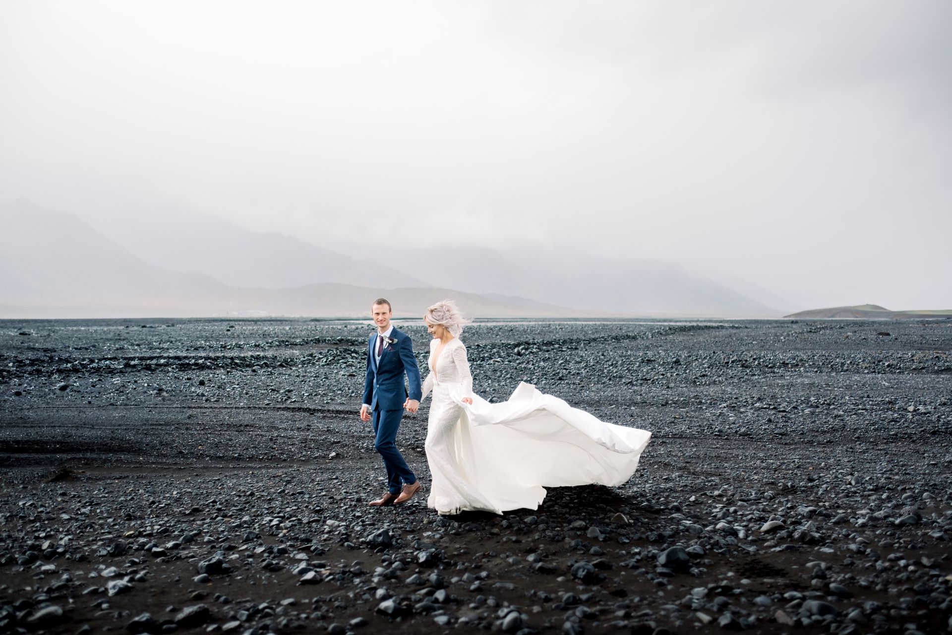 A bride and groom are walking on a rocky beach.