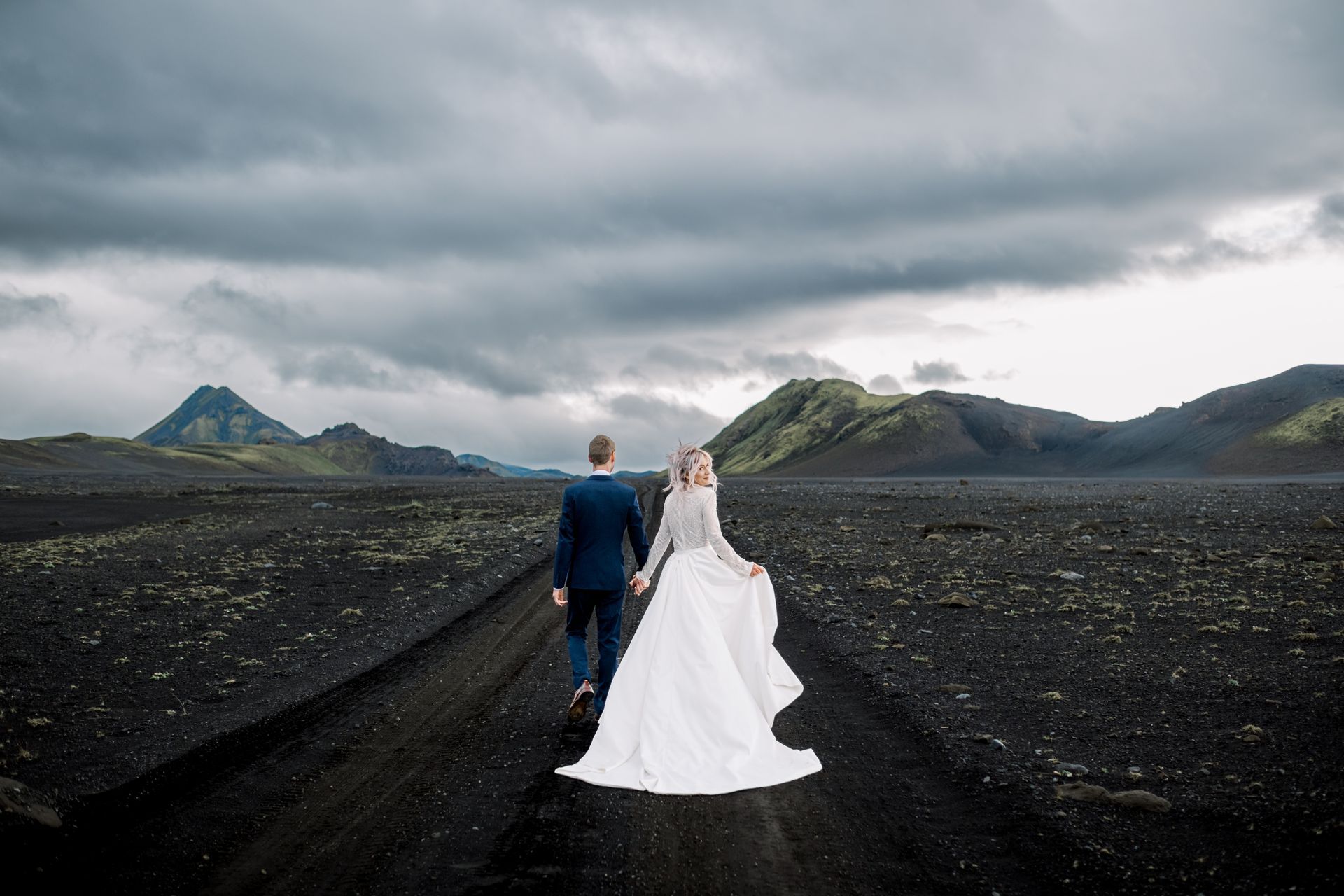 A bride and groom are walking down a dirt road holding hands.
