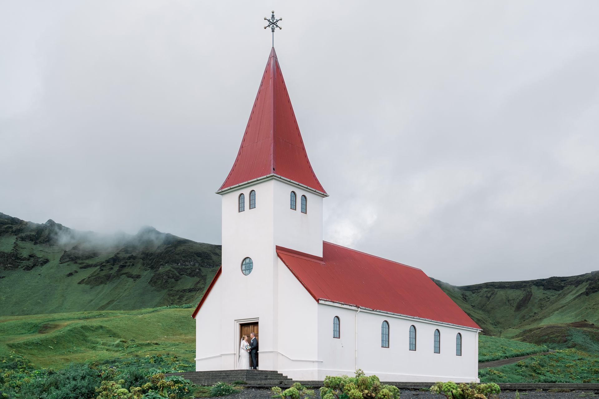 A newlywed couple poses in front of a white church with a red roof in Iceland.