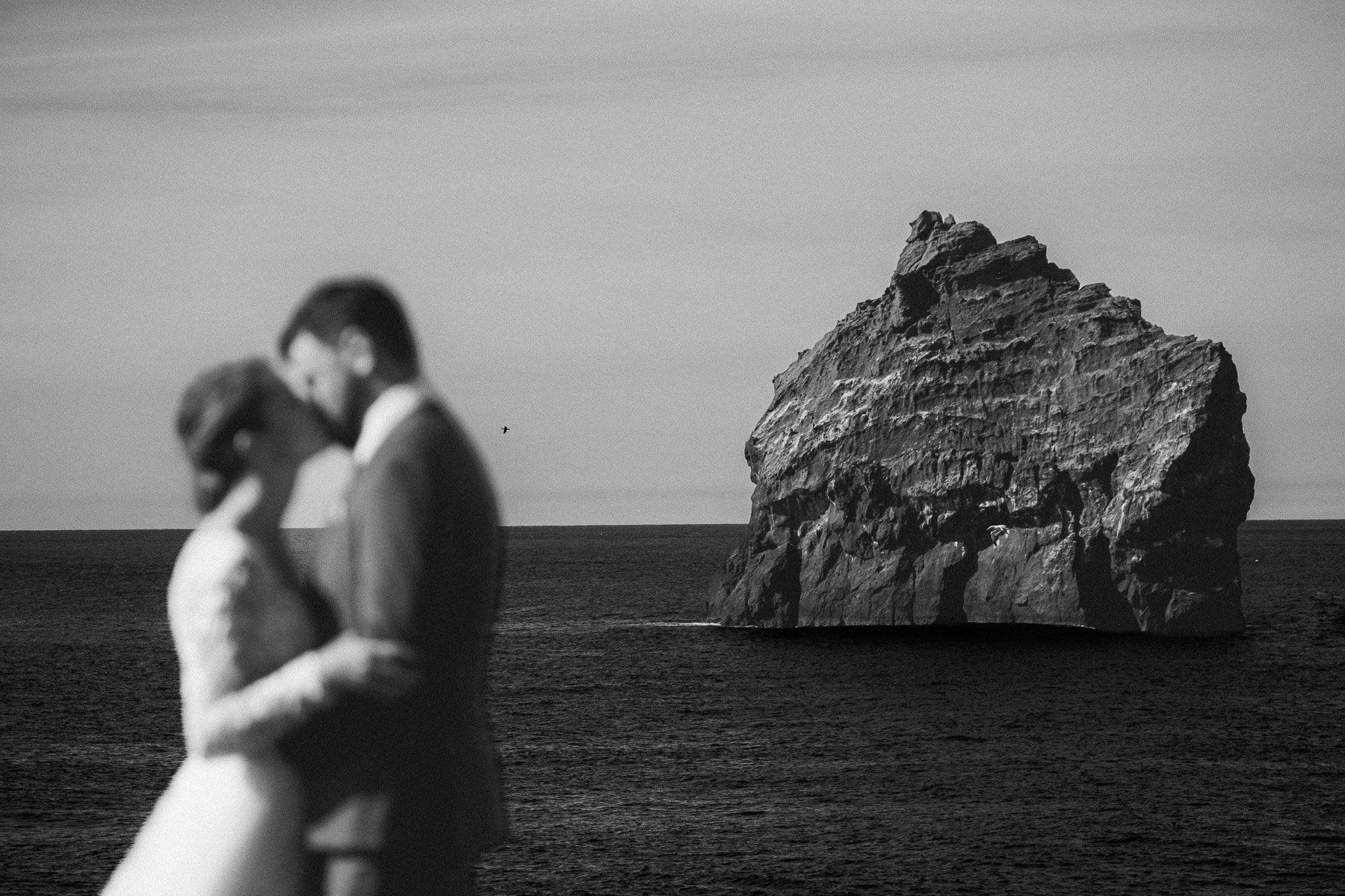 A bride and groom are dancing in front of a large rock in the ocean.
