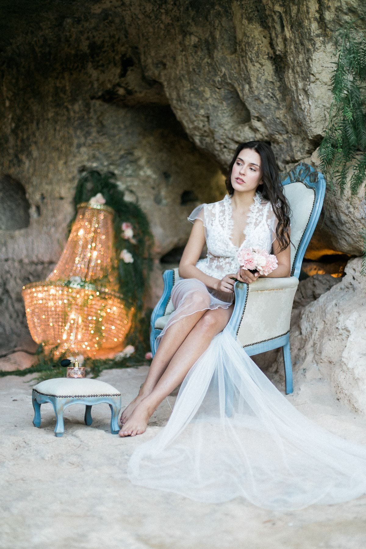A woman in a wedding dress is sitting in a chair in a cave holding a bouquet of flowers.