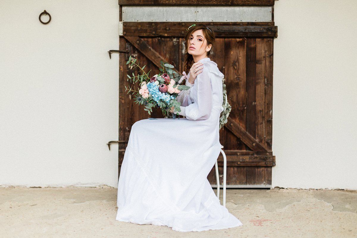 A woman in a white dress is sitting on a chair in front of a wooden door holding a bouquet of flowers.