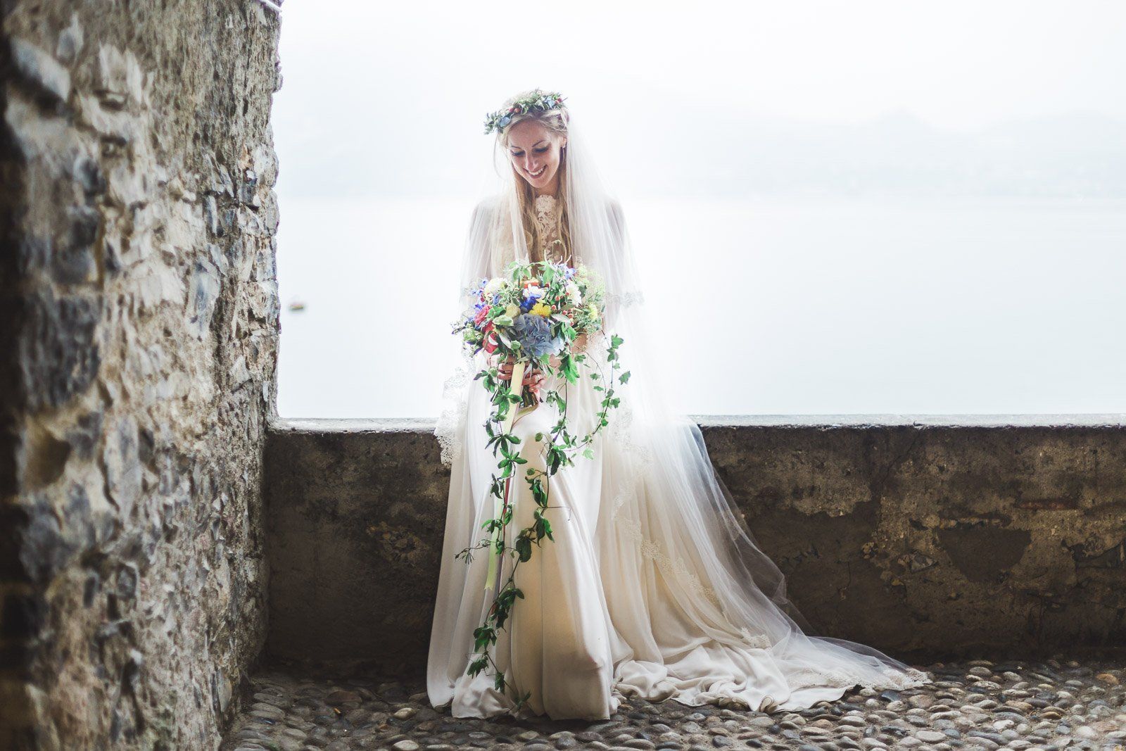 A bride in a wedding dress is standing next to a stone wall holding a bouquet of flowers.