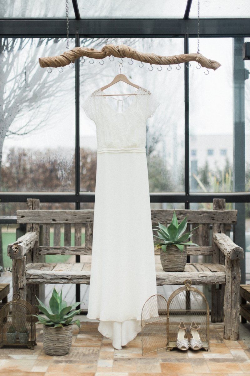 A wedding dress is hanging on a wooden bench in a greenhouse.