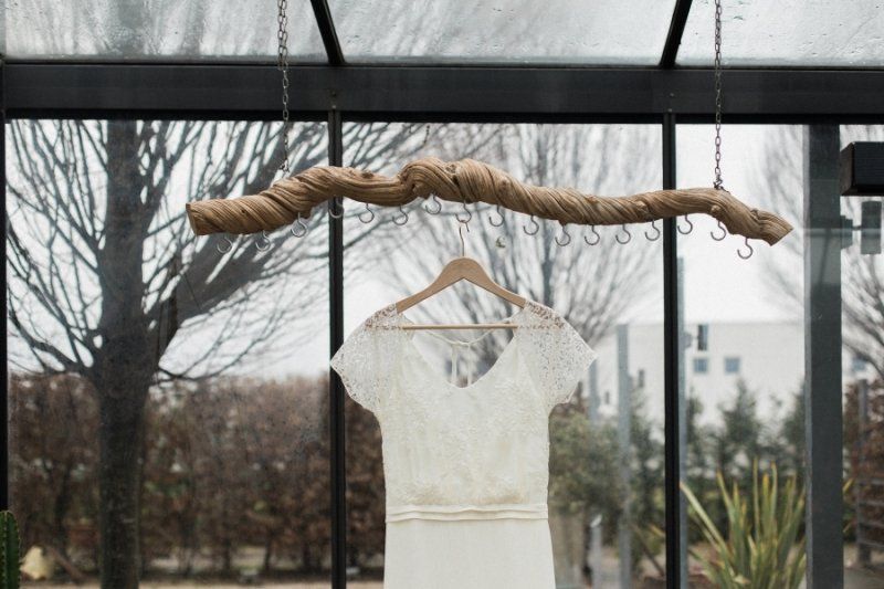 A wedding dress is hanging on a wooden hanger in a greenhouse.