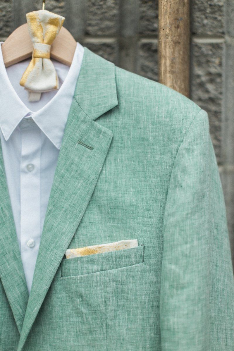 A green suit jacket with a yellow bow tie is hanging on a wooden hanger.