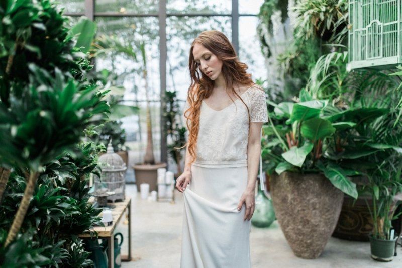 A woman in a white dress is standing in a greenhouse surrounded by plants.