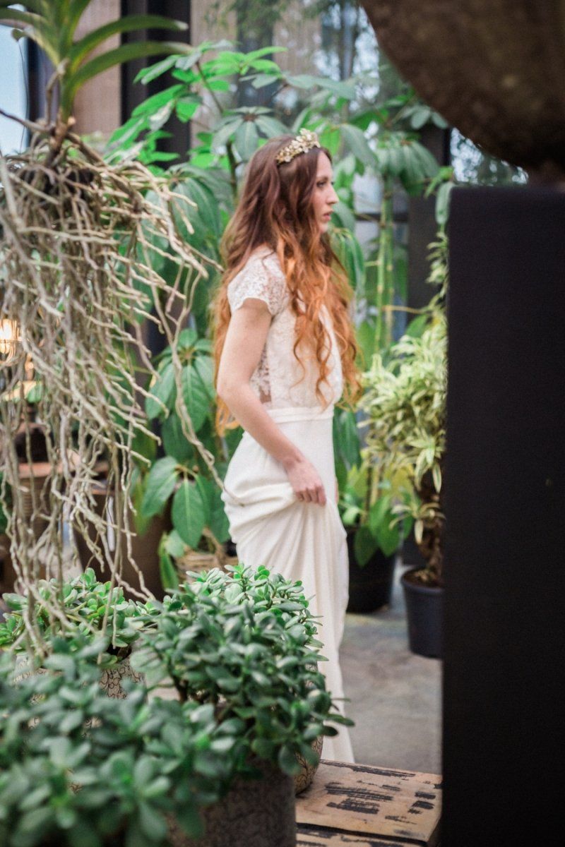 A woman in a white dress is standing in a greenhouse surrounded by plants.