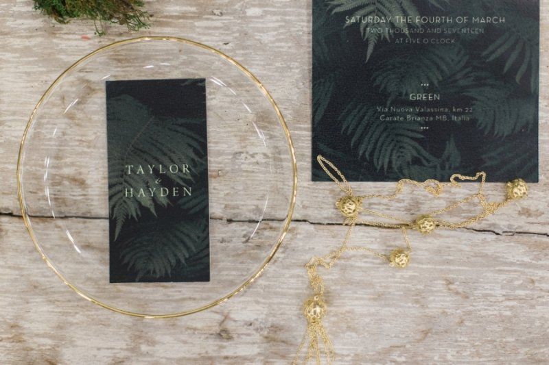 A black and gold wedding invitation is sitting on a wooden table next to a glass plate.