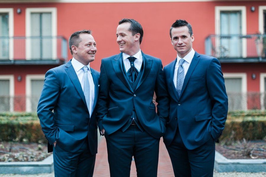 Three men in suits and ties are standing next to each other in front of a red building.