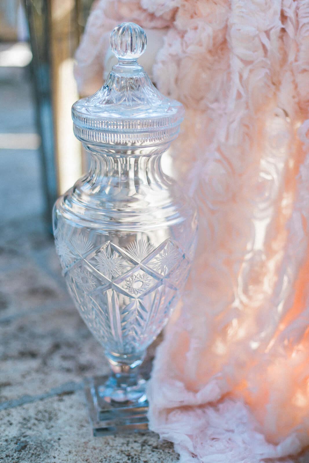 A clear glass vase with a lid is sitting on a table next to a pink blanket.