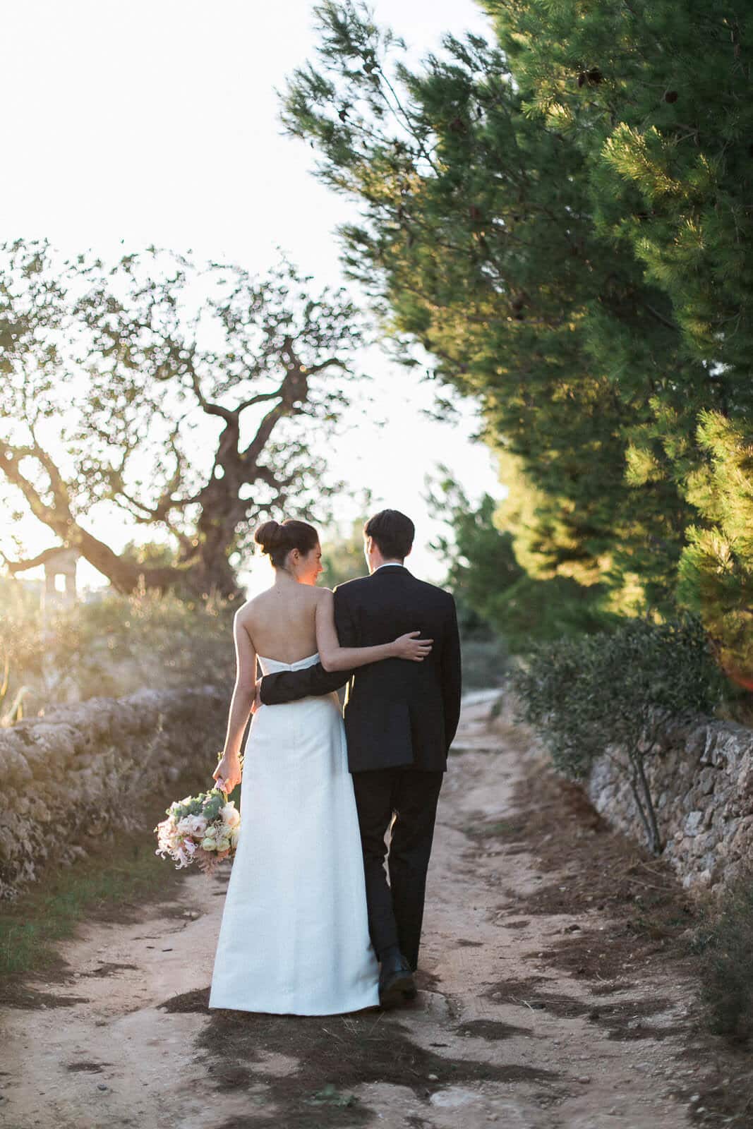 A bride and groom are walking down a dirt road.