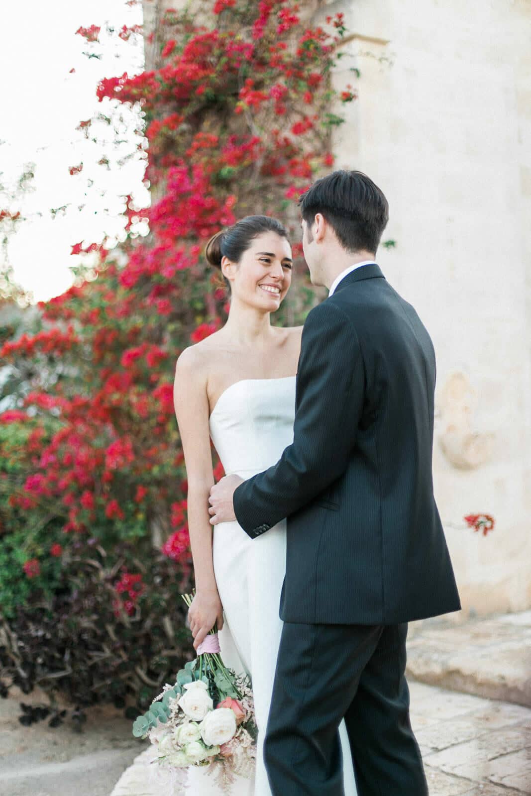 A bride and groom are standing next to each other in front of a bush with red flowers.