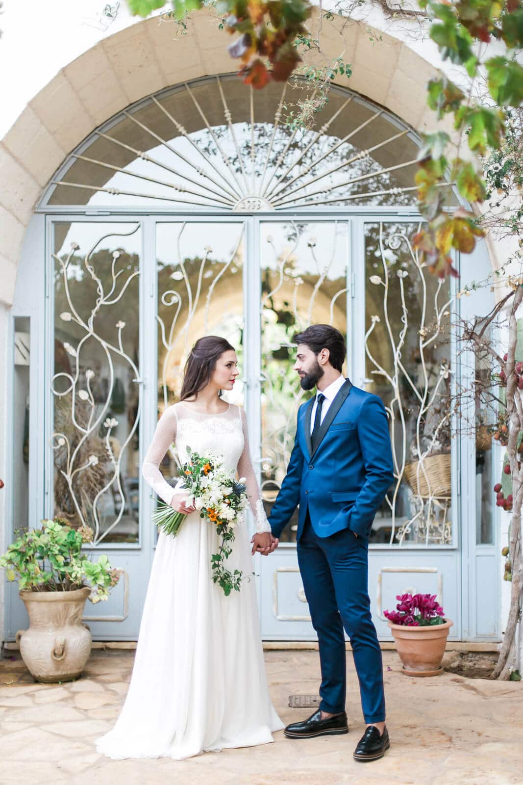 A bride and groom are standing in front of a building holding hands.