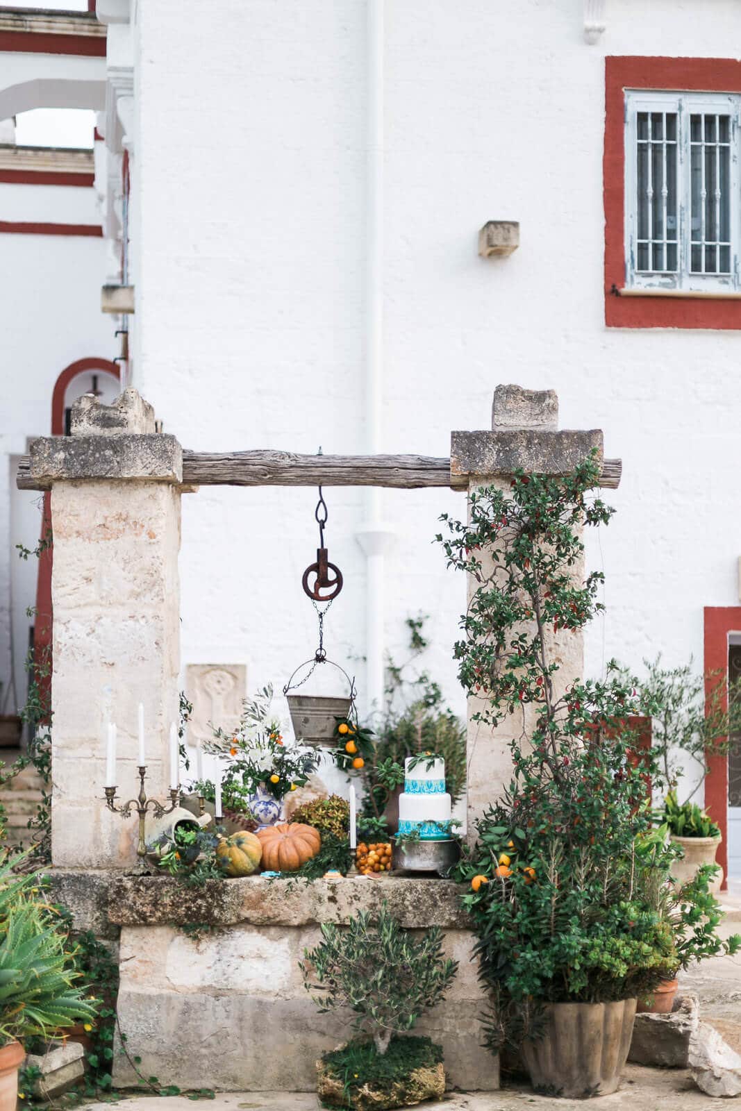 A stone well surrounded by plants and pumpkins in front of a white building.
