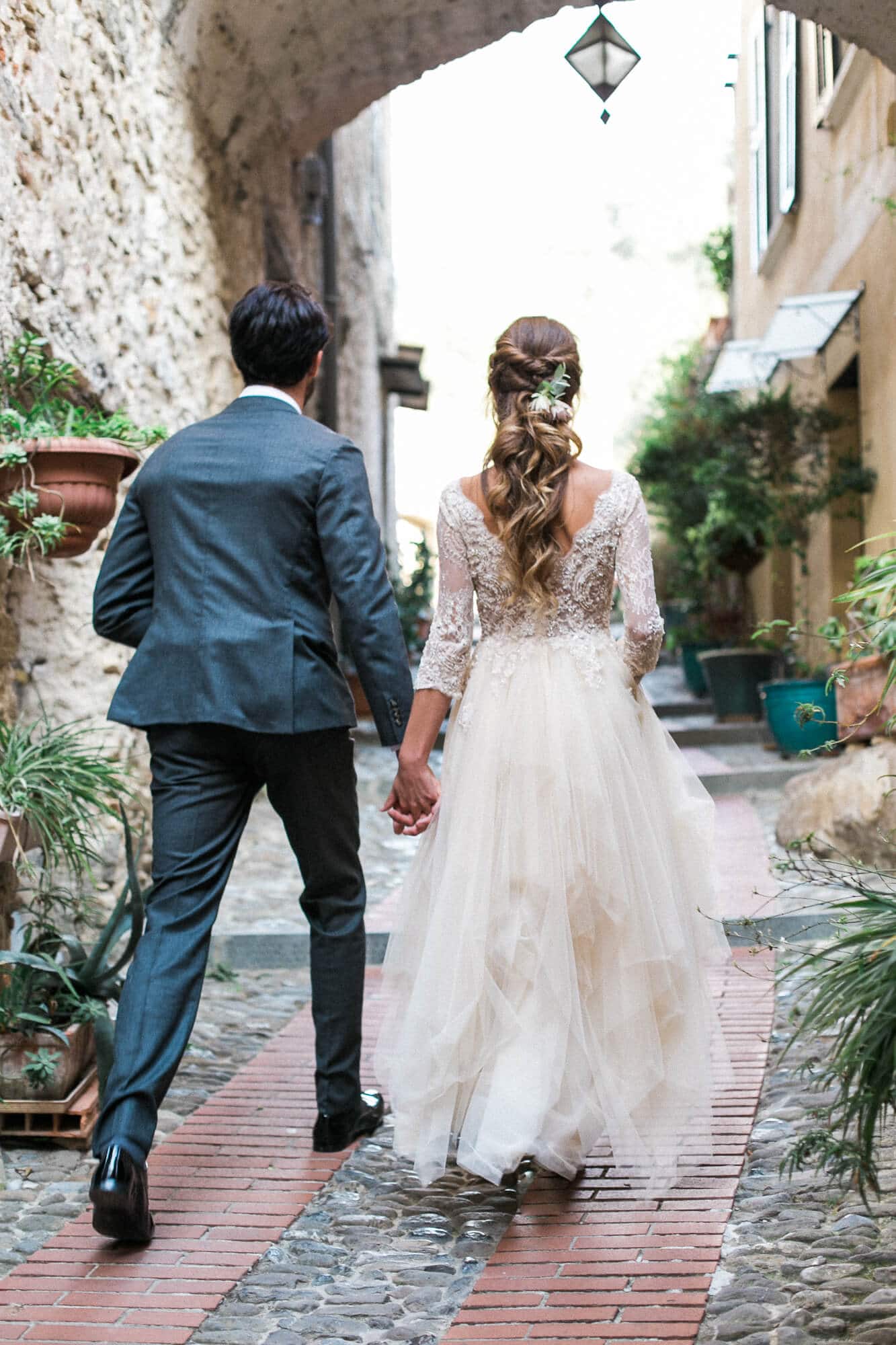 A bride and groom are walking down a narrow street holding hands.