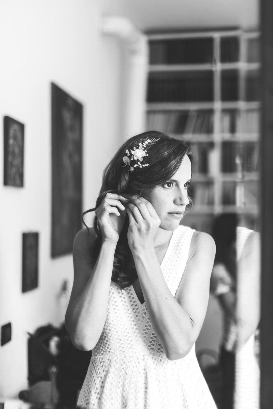 A woman in a white dress is putting on her earrings in front of a mirror.