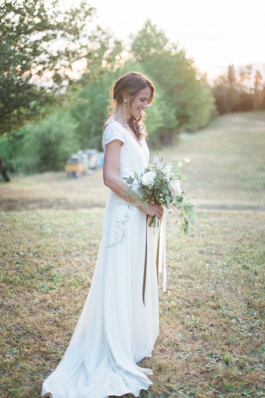 A bride in a white dress is standing in a field holding a bouquet of flowers.