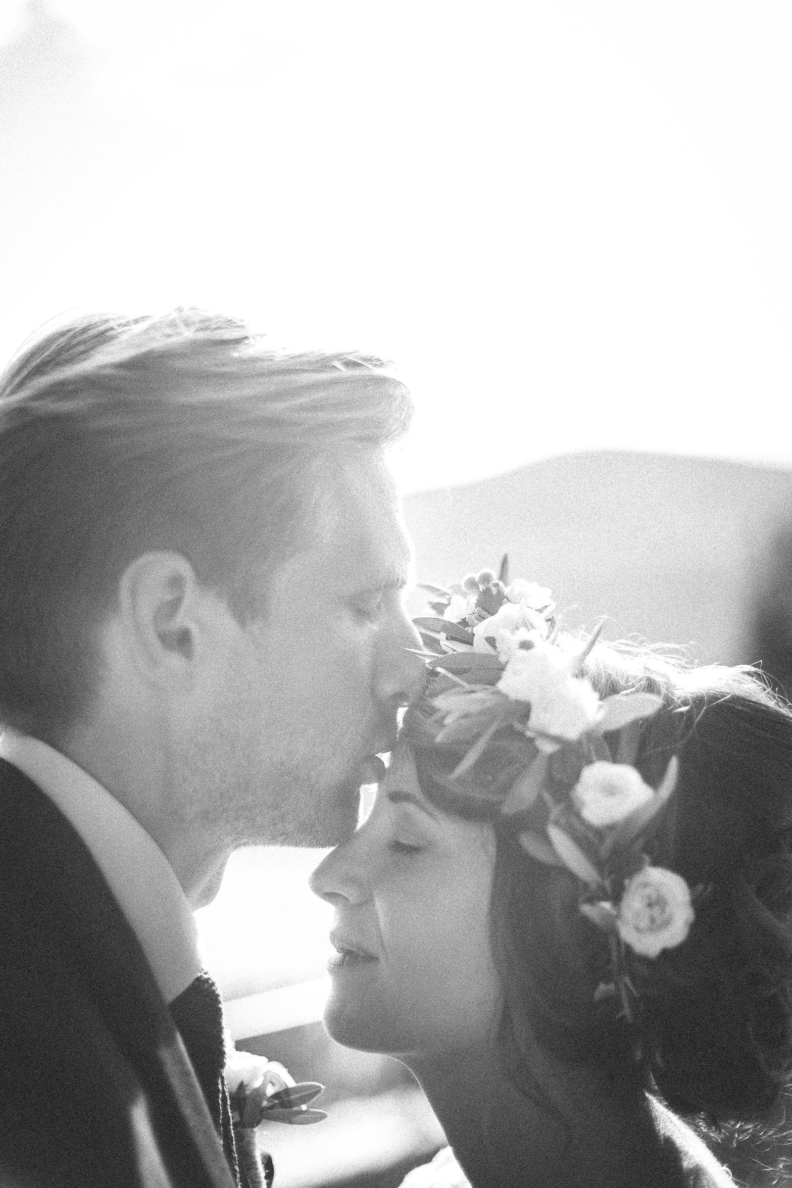 A bride and groom are kissing in a black and white photo . the bride has a flower crown on her head.