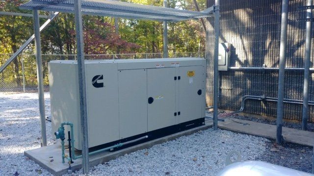 A large generator is sitting under a fence in a gravel area.