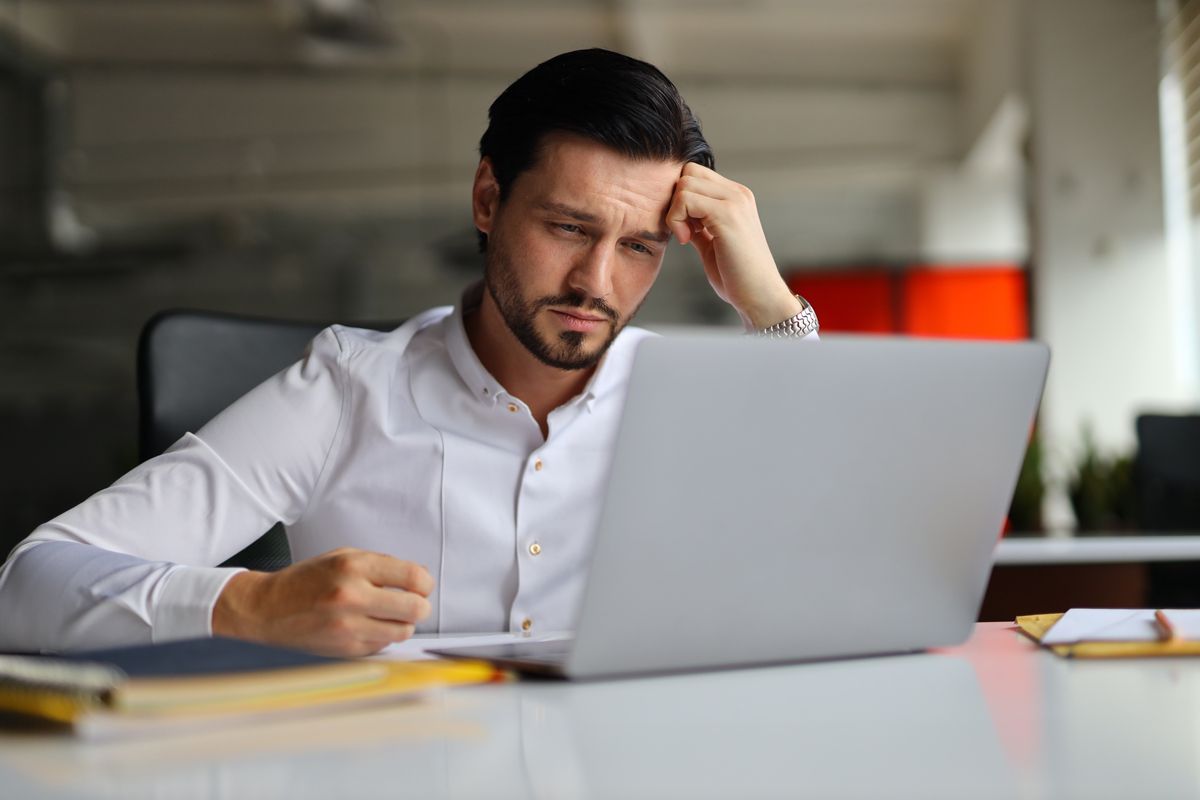 Man sitting at a desk, looking stressed while working on a laptop in an office