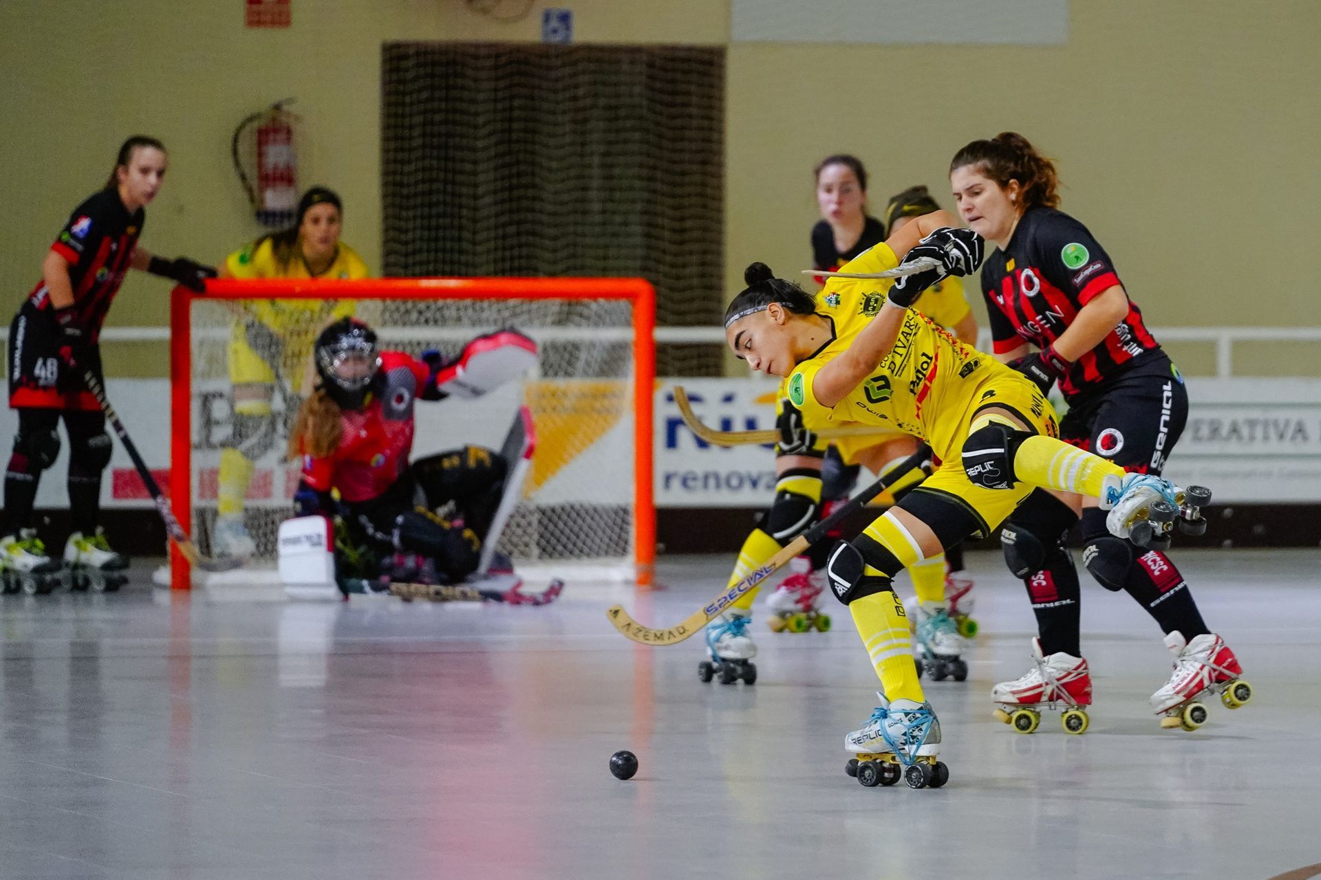 Partido de hockey sobre patines: un jugador de amarillo dispara, defendido por un jugador de rojo. El portero está en la portería.