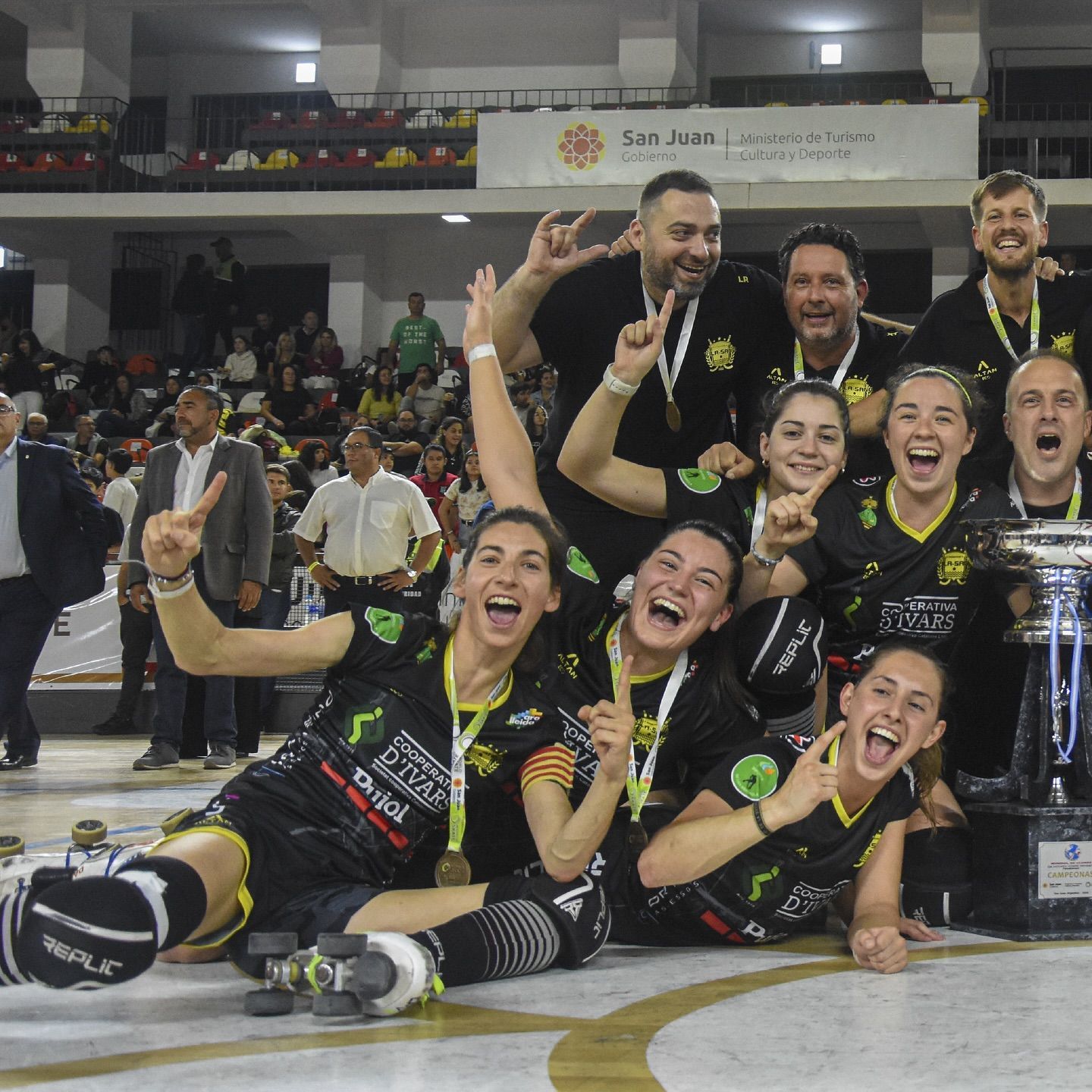 El equipo de hockey sobre patines celebra la victoria, vestidos de negro y amarillo, posando con el trofeo, en el interior.