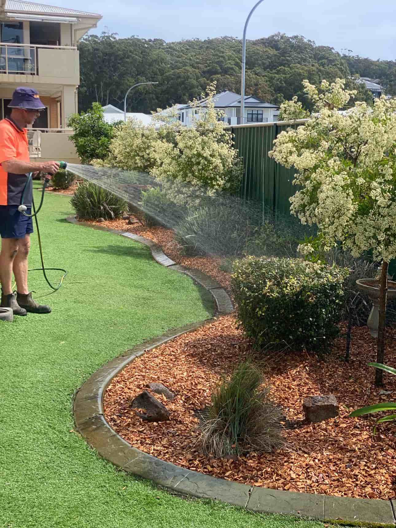 A Man Is Watering A Lawn With A Hose — Gibbo’s Lawns & Property Maintenance In Salamander Bay, NSW