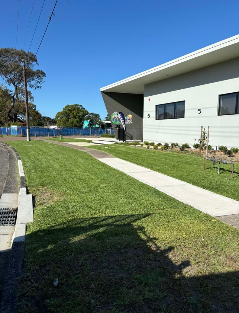 A Large White Building With A Walkway Leading To It Is Surrounded By Grass And Trees — Gibbo’s Lawns & Property Maintenance In Salamander Bay, NSW