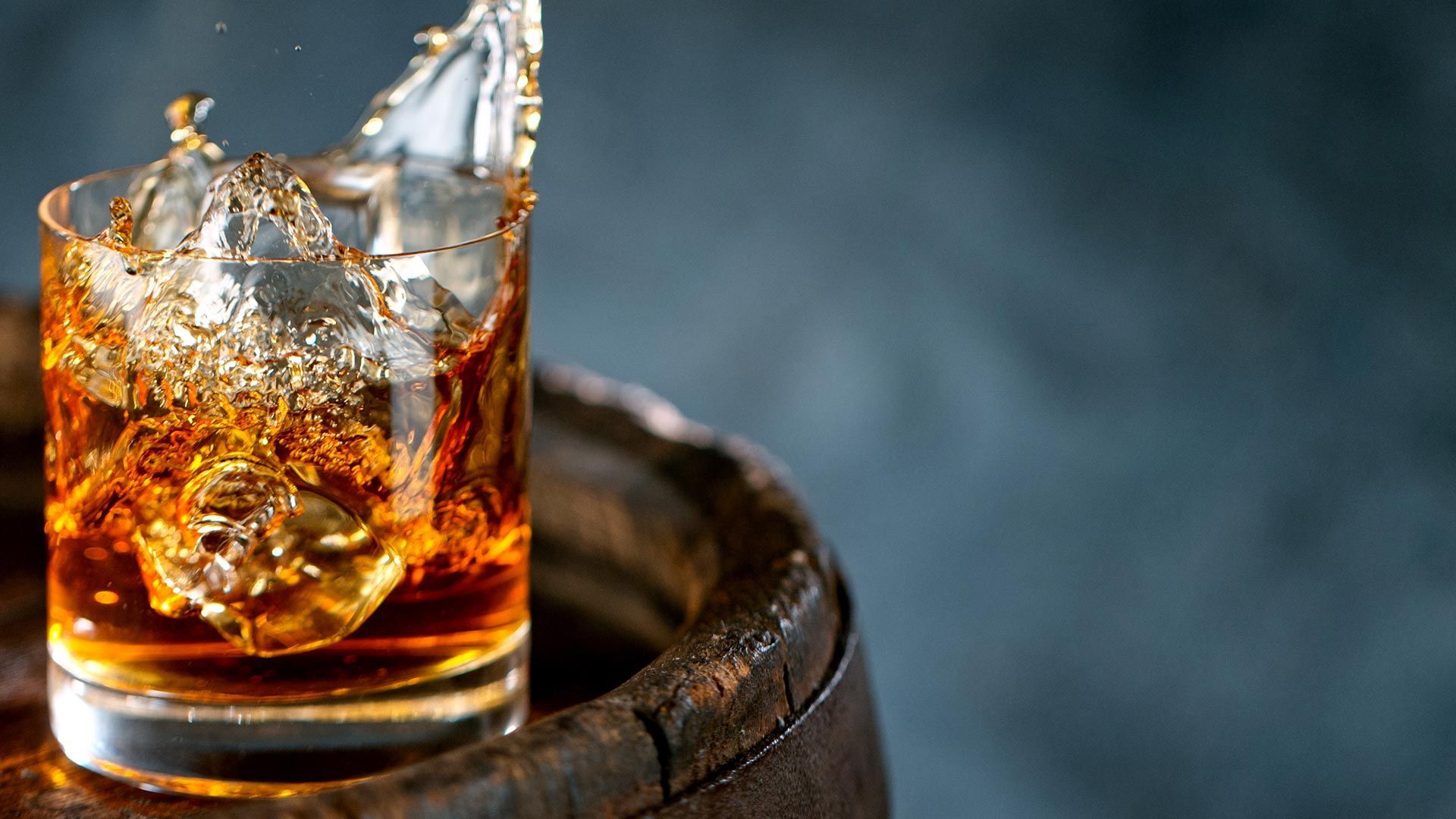 A glass of whiskey with ice is sitting on top of a wooden barrel.