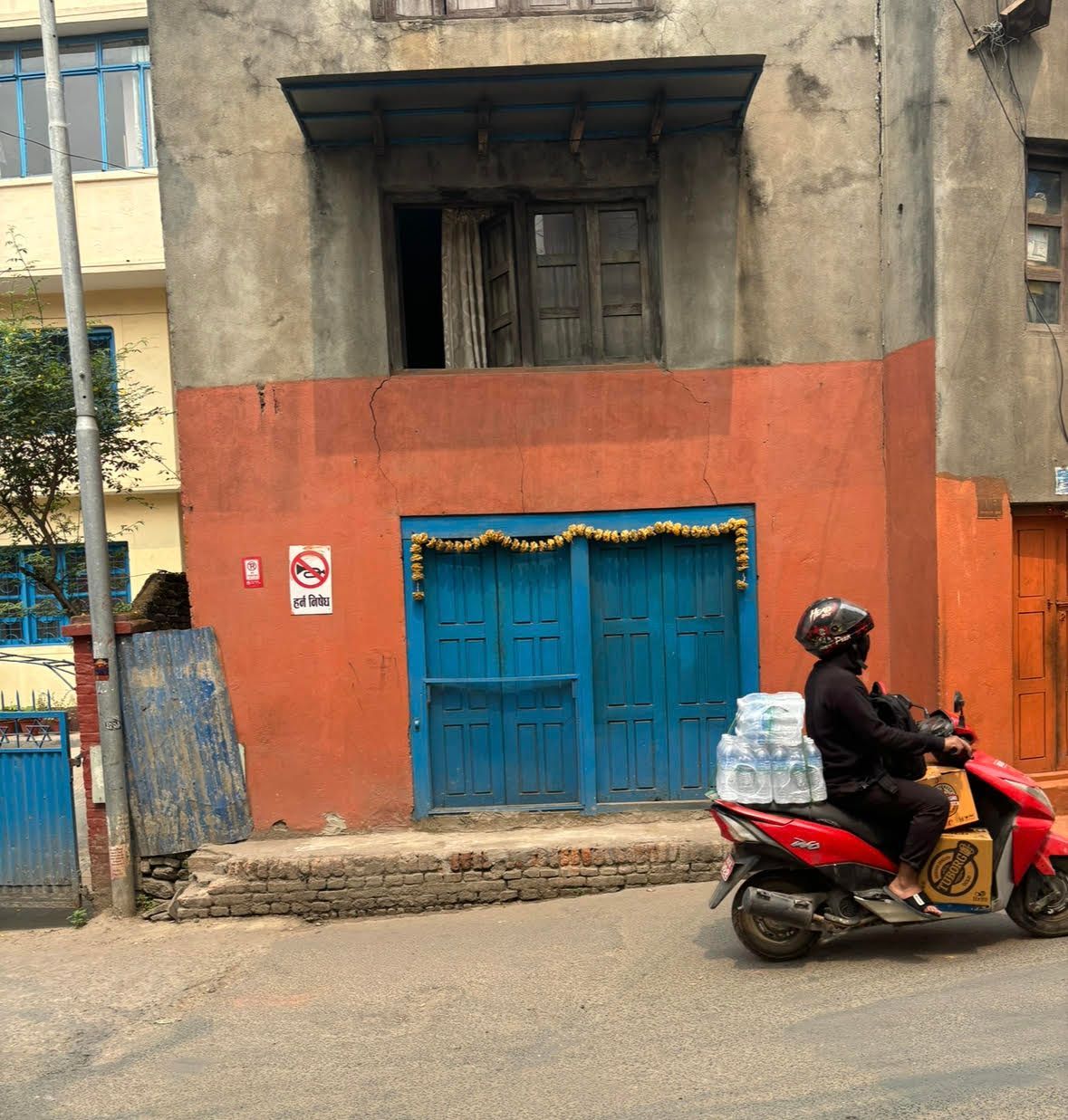 A person on a red scooter delivers water in front of a blue-doored building with an orange facade.