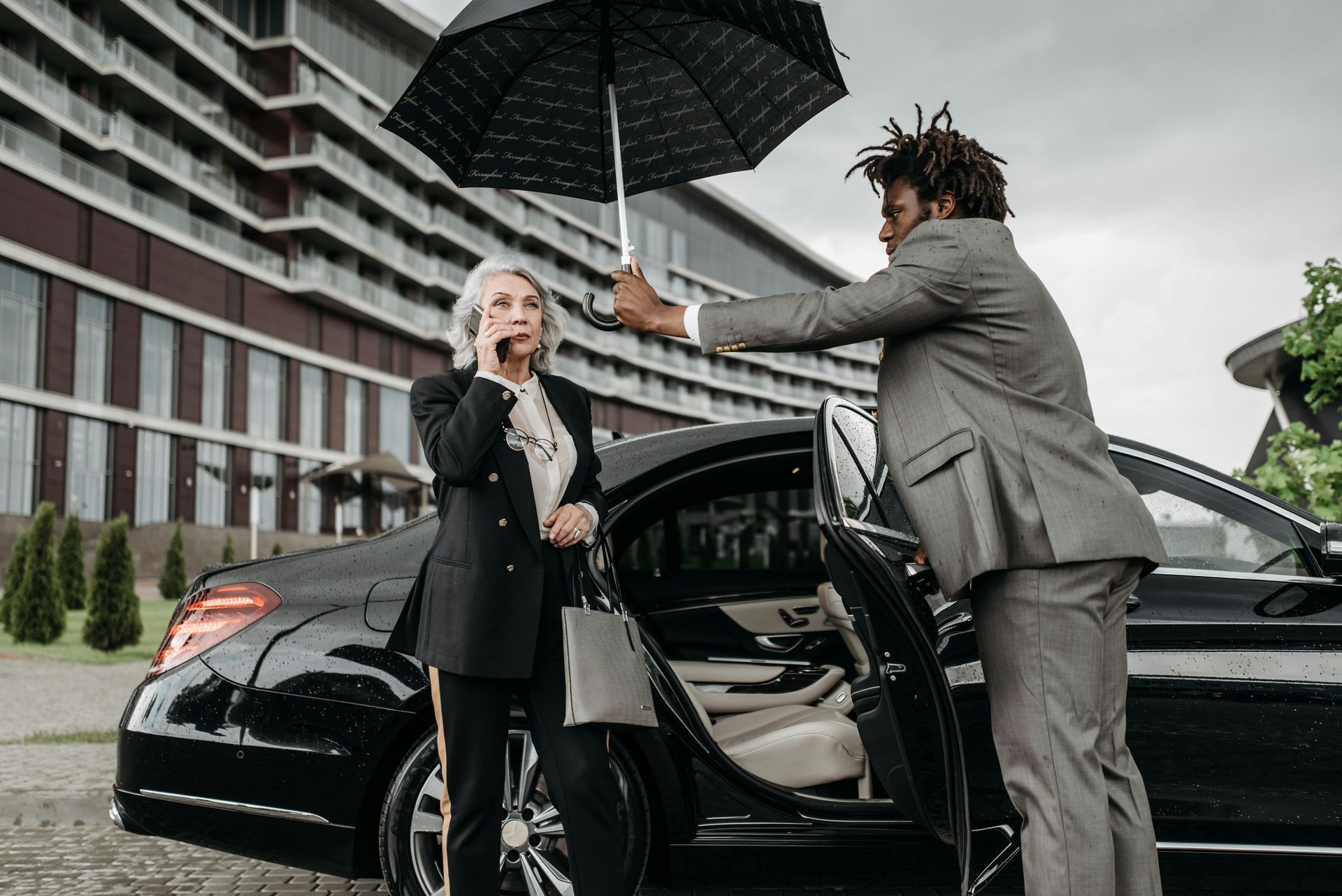 Man holding umbrella for person exiting black car in front of a building; person on phone.
