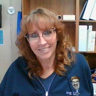 A woman wearing glasses and a blue shirt is smiling in front of a bookshelf.