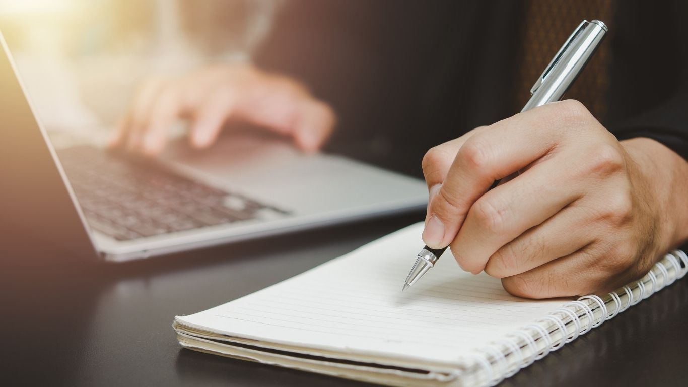 Person writing notes beside a laptop, symbolising the detailed review and analytical process involve