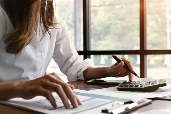 Woman using a calculator and reviewing documents at a desk in a well-lit office.