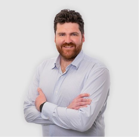 Man with brown hair and beard, wearing a light blue button-down shirt, arms crossed, smiling.