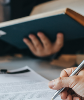 Hand with pen signing document; book open in background.