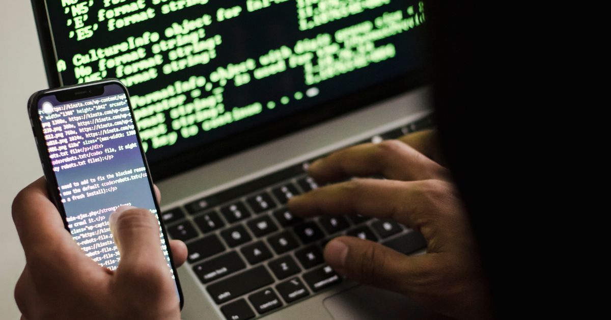 Hands using a smartphone and laptop displaying code, symbolising cybercrime investigation and digita