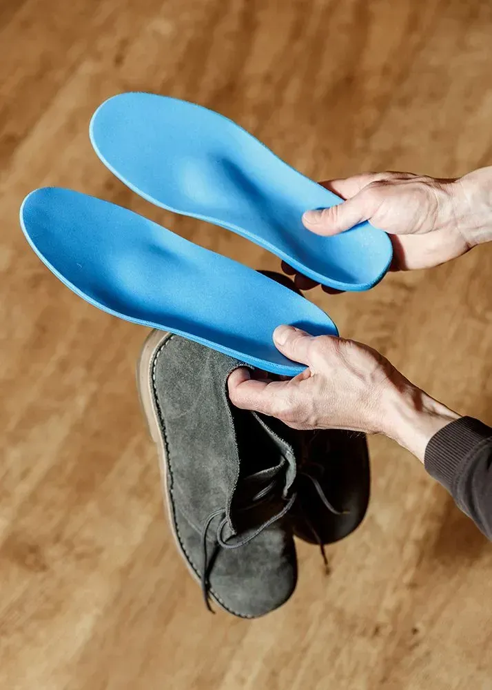 A person holding two blue orthopedic insoles above a grey suede desert boot against a wooden background.