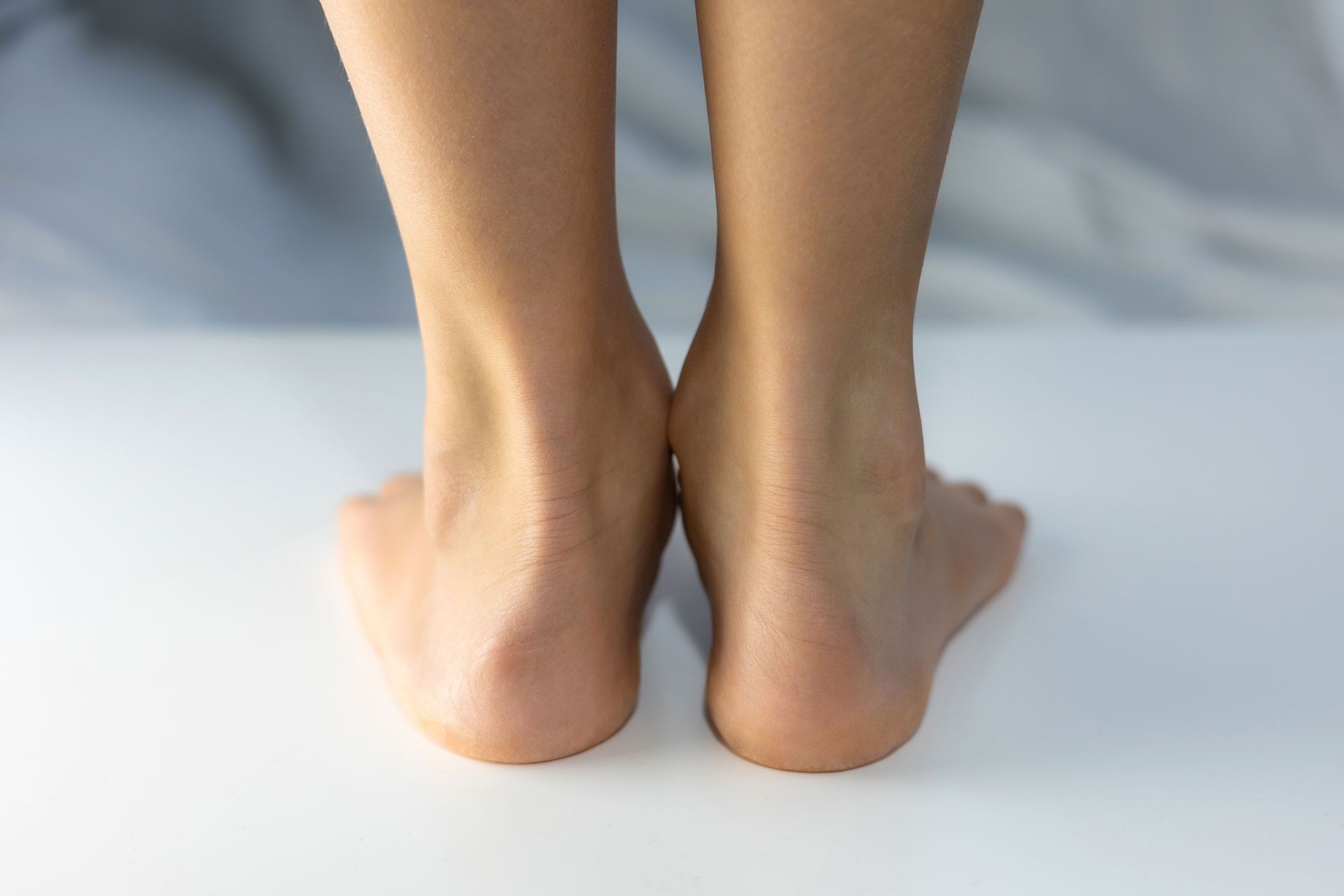 Close-up, rear view of two bare feet standing on a flat, white surface, showing slight inward tilt of the heels.