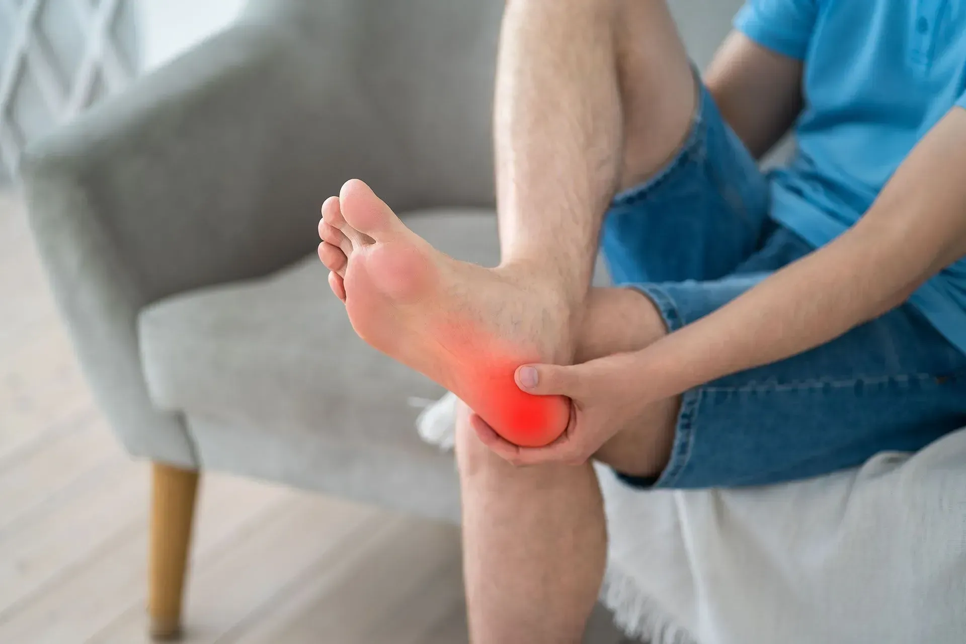 A person sitting in a chair, holding their foot with a red highlighted area indicating pain in the heel and arch.