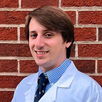 A person in a white lab coat, blue striped shirt, and patterned tie smiles in front of a red brick wall.
