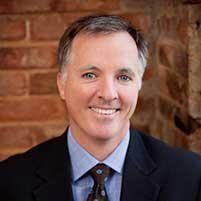 Headshot of a smiling person in a blue shirt and dark suit jacket, positioned against a rustic brick wall background.